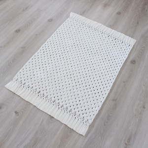 May include: A white macrame rug with a diamond pattern and fringe edges. The rug is made of cotton cord and is perfect for adding a touch of bohemian style to any room.