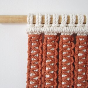 Puede incluir: Un tapiz de macram&eacute; de pared con una barra de madera en la parte superior. El tapiz est&aacute; hecho con cord&oacute;n blanco y naranja y tiene un patr&oacute;n geom&eacute;trico.