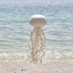 Peut inclure: Suspension murale en macramé en forme de méduse blanche, avec une tête ronde et de longs tentacules en spirale. L'œuvre est suspendue devant un arrière-plan flou de l'océan, avec la mer et le ciel visibles.