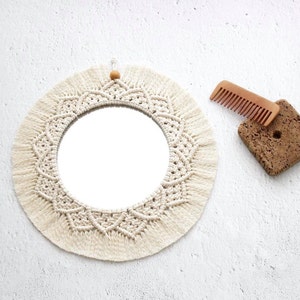 May include: A round mirror with a white macrame frame and fringe. The mirror is hanging on a white wall.