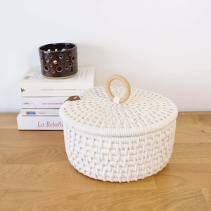 May include: White woven storage basket with a lid and a wooden handle. The basket is sitting on a wooden surface.