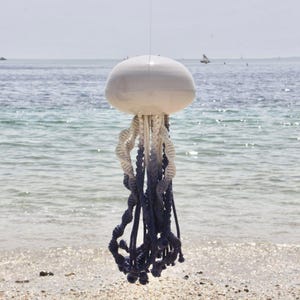 May include: A decorative jellyfish sculpture with a white, rounded top and trailing tentacles. The tentacles transition from white to a deep navy blue. The sculpture is suspended in front of a beach and ocean backdrop.