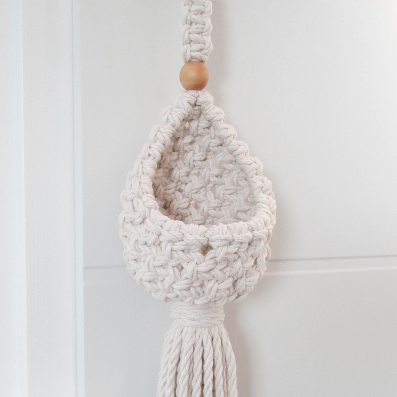 May include: A white macrame hanging planter with a wooden bead and a long tassel. The planter is made of cotton cord and has a woven design.