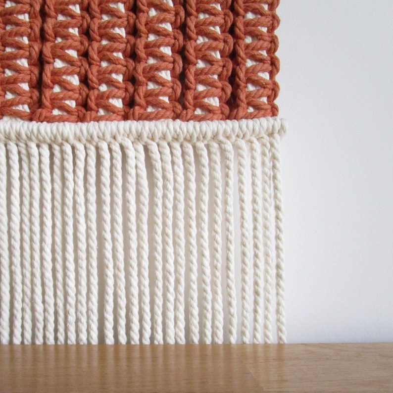 Puede incluir: Un tapiz de macram&eacute; de pared con una secci&oacute;n tejida de color terracota y una franja blanca.