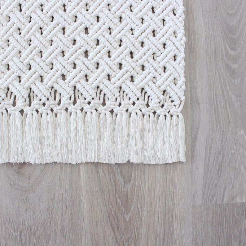 May include: A white macrame rug with a fringe border. The rug has a diamond pattern and is made of cotton cord.