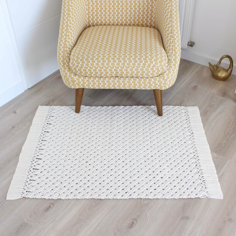May include: A white macrame rug with fringe edges, placed on a light wood floor, in front of a yellow patterned armchair.