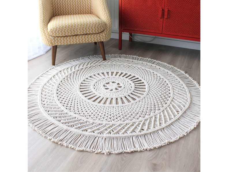 Peut inclure: Un tapis rond en macram&eacute; blanc avec une bordure en franges. Le tapis a un motif circulaire au centre et un design g&eacute;om&eacute;trique autour du bord.