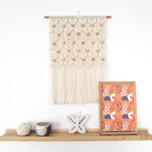 May include: A white macrame wall hanging with wooden beads, a framed print with a colourful abstract pattern, a small ceramic bowl, a ball of yarn, and a white butterfly decoration on a wooden shelf.