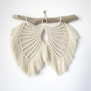 May include: A white macrame wall hanging with a fan shape and fringe. The hanging is attached to a piece of driftwood.