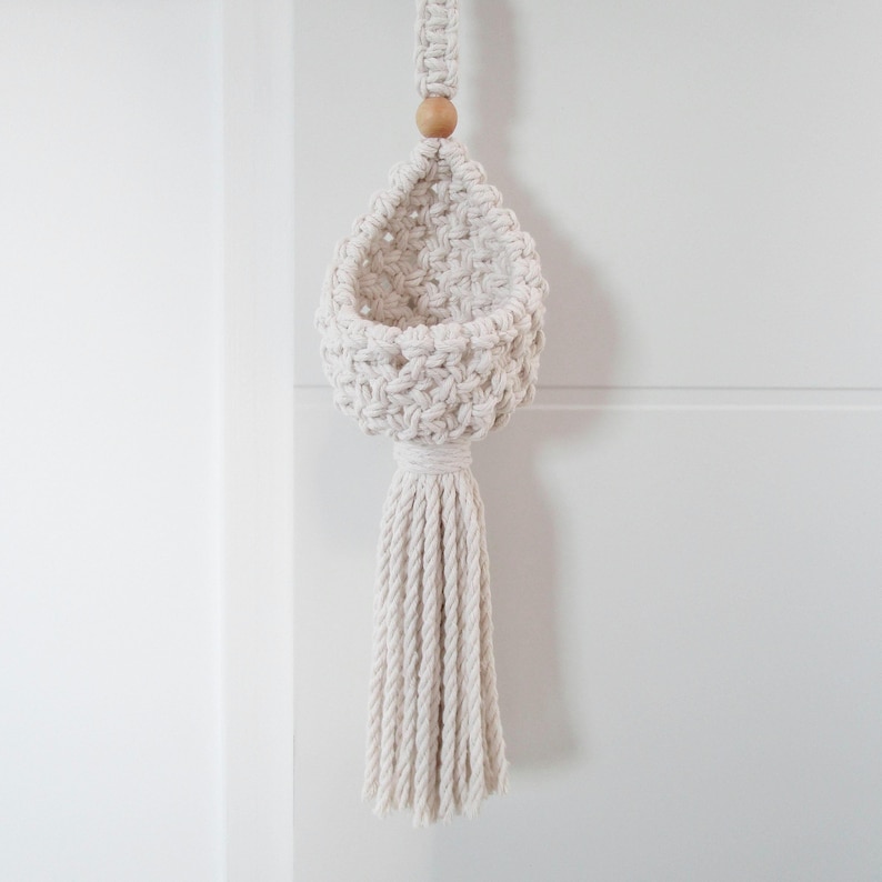 May include: A white macrame hanging planter with a wooden bead at the top and a long tassel. The planter is empty and ready to hold a small plant.