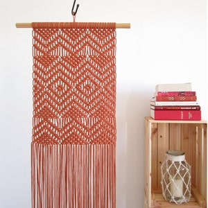 May include: A terracotta-coloured macrame wall hanging with a geometric pattern. The hanging has a wooden dowel rod at the top and long fringe at the bottom.