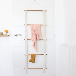 Op de afbeelding: Witte macramé wandhanger met drie houten latten en een roze handdoek en een geel hoofdband die aan de latten hangen.