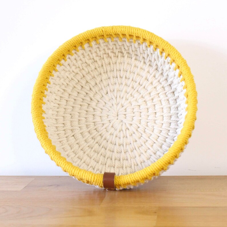 Corbeille de rangement, réalisée avec de la corde écrue et jaune, posée contre un mur blanc