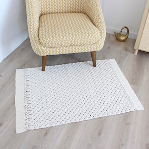 Peut inclure: Un tapis tissé blanc avec un motif en losange et des franges sur les bords. Le tapis est placé sur un sol en bois clair et une chaise à motifs jaunes est posée dessus.