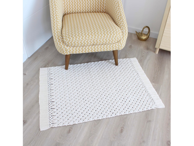 May include: A white woven rug with a diamond pattern and fringe edges. The rug is placed on a light wood floor and a yellow patterned chair is sitting on top of it.