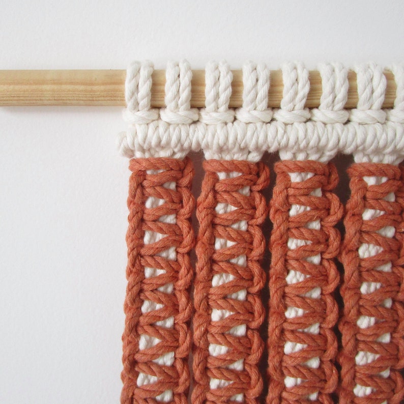 Puede incluir: Un tapiz de macram&eacute; de pared con una barra de madera en la parte superior. El tapiz est&aacute; hecho con cord&oacute;n blanco y naranja y tiene un patr&oacute;n geom&eacute;trico.