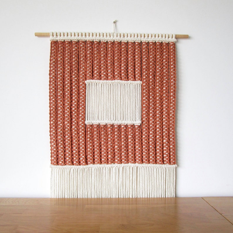 Puede incluir: Un tapiz de macram&eacute; de pared con un dise&ntilde;o cuadrado. El tapiz est&aacute; hecho con cord&oacute;n de color natural y presenta un cuadrado blanco en el centro. El tapiz tiene una franja en la parte inferior.