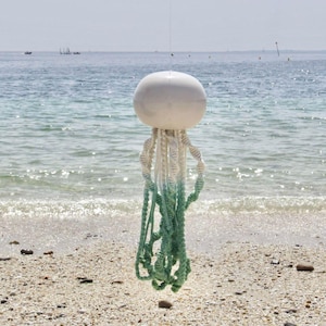 May include: A white, spherical buoy with trailing, twisted rope tentacles in shades of white and green. The decorative piece hangs against a backdrop of a sandy beach and ocean, with boats visible in the distance.