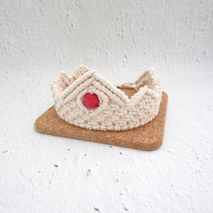 May include: A white macrame crown with a red bead in the centre. The crown is sitting on a cork coaster.