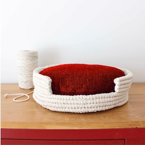 May include: A white rope pet bed with a red fabric cushion inside. The bed is on a wooden surface.