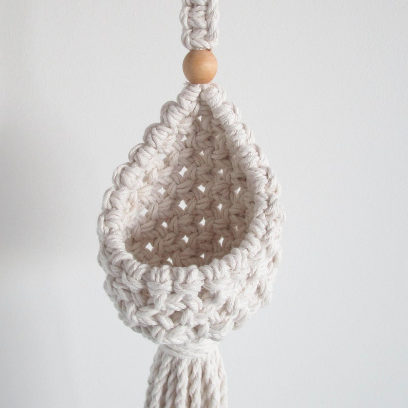 May include: A white macrame hanging planter with a wooden bead at the top. The planter is empty and has a tassel at the bottom.