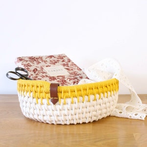 May include: A white and yellow crocheted basket with a brown leather handle. The basket is filled with a book, scissors, and white lace.