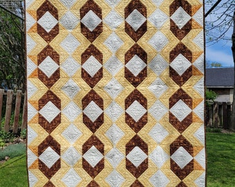 Pode incluir: Uma colcha com um padrão geométrico em tons de marrom, amarelo e branco. A colcha está pendurada em um varal ao ar livre.