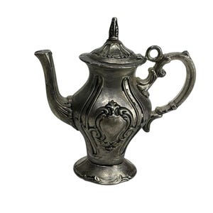 May include: A silver-toned metal teapot featuring an ornate, embossed scrollwork pattern on the body, a curved handle, a pointed lid with a finial, and a flared base. The surface has a weathered, metallic finish with visible dark patina.