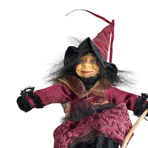 May include: A Halloween witch doll with a red and black color scheme. The witch has a pointed hat, black hair, and a smiling face. Her outfit includes a red dress with lace accents and black gloves. A decorative item for Halloween.