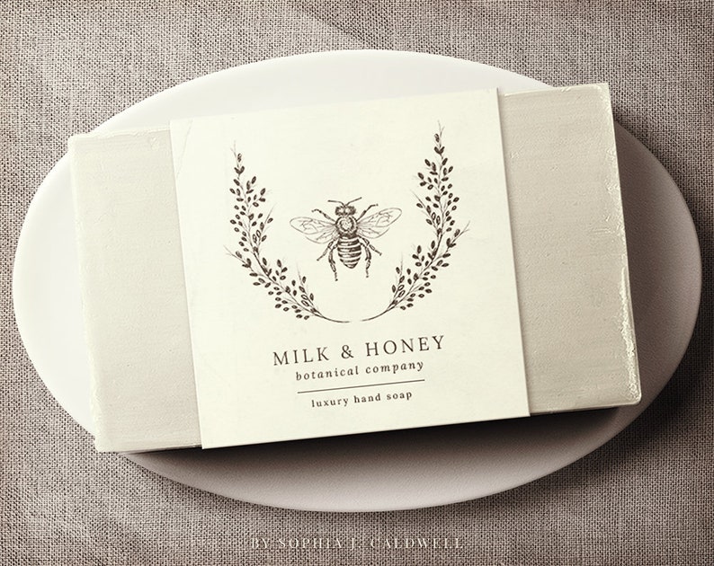 May include: A white bar of soap with a label that reads "Milk & Honey botanical company luxury hand soap" and features a black and white illustration of a bee inside a wreath of leaves.