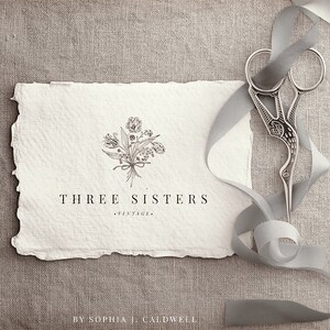 May include: A white card with a hand-drawn floral design and the text "THREE SISTERS - VINTAGE - BY SOPHIA J. CALDWELL". A silver pair of scissors with a decorative design is on a gray ribbon.