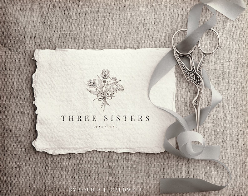 May include: A white card with a hand-drawn floral design and the text "THREE SISTERS - VINTAGE - BY SOPHIA J. CALDWELL". A silver pair of scissors with a decorative design is on a gray ribbon.