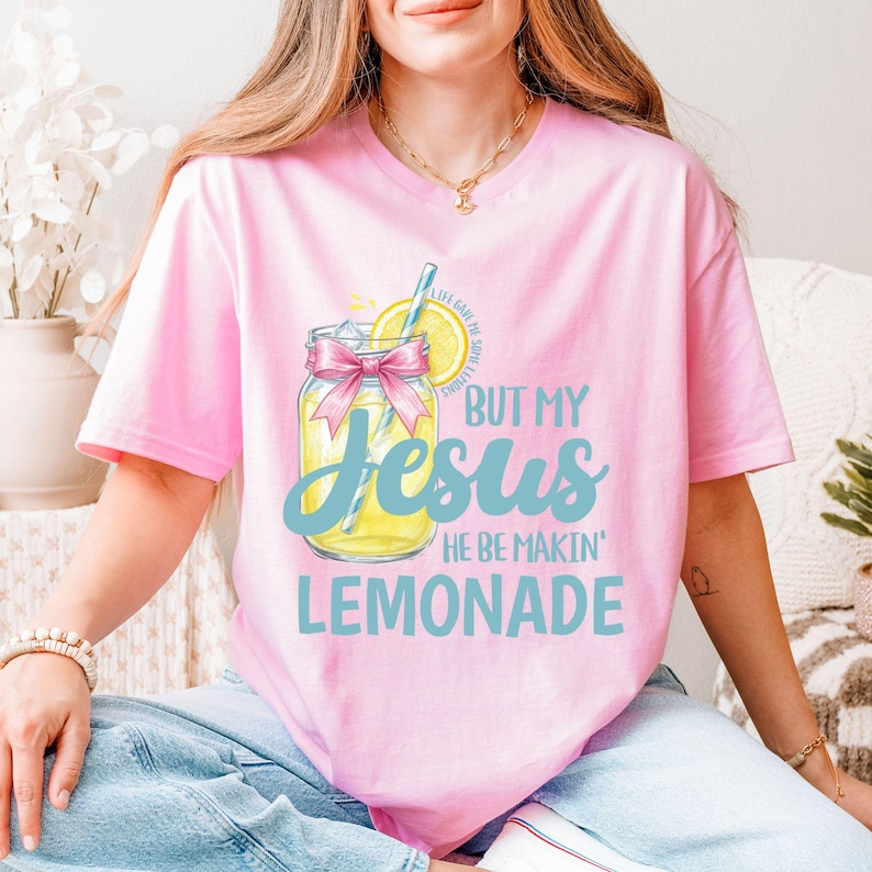 May include: Pink t-shirt with a lemonade graphic. The graphic features a mason jar with a pink bow, a lemon slice, and the text "BUT MY Jesus HE BE MAKIN' LEMONADE" in blue and yellow.