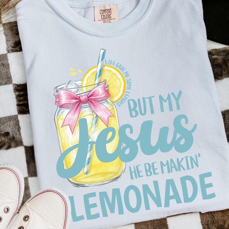 May include: Light blue t-shirt with a lemonade graphic. The graphic features a mason jar with a pink bow, a lemon slice, and a straw. Text on the shirt reads "BUT MY Jesus HE BE MAKIN' LEMONADE".