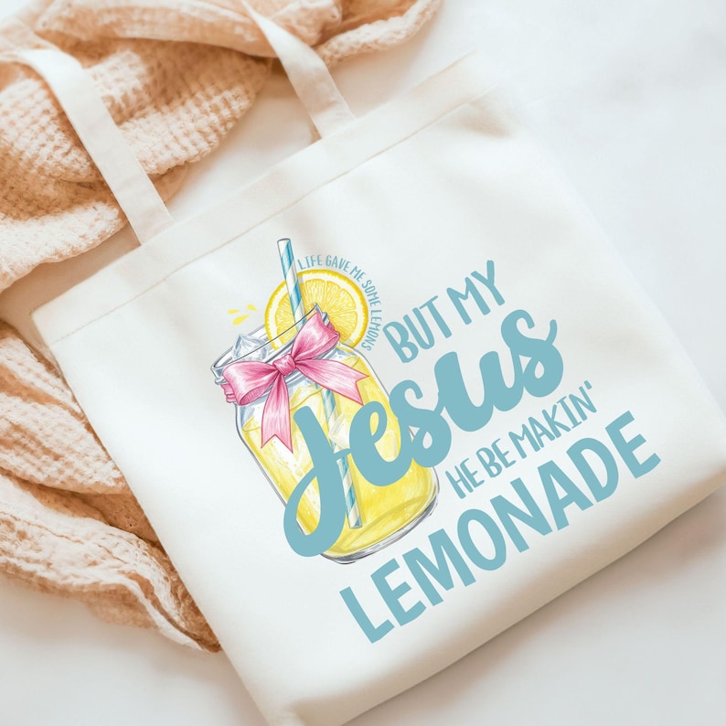 May include: White tote bag with a graphic of a lemonade drink with a pink bow and a lemon slice. Text on the bag reads "But My Jesus He Be Makin' Lemonade" in blue script. The phrase "Life Gave Me Some Lemons" is also printed on the bag.