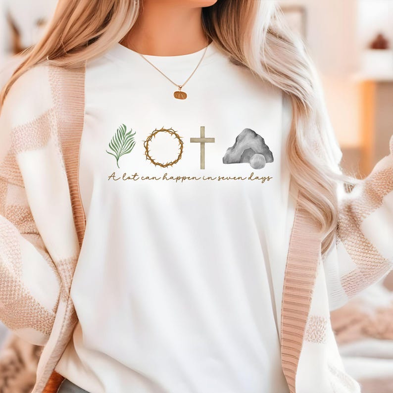 May include: White sweatshirt with a watercolor design featuring a palm branch, crown of thorns, cross, and empty tomb. The text "A lot can happen in seven days" is printed below the images. The design is in shades of green, brown, and gray.