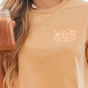 Puede incluir: Una camiseta de color beige con las palabras "MADE for more" en blanco. Las palabras est&aacute;n rodeadas de un patr&oacute;n a cuadros en naranja y rosa.