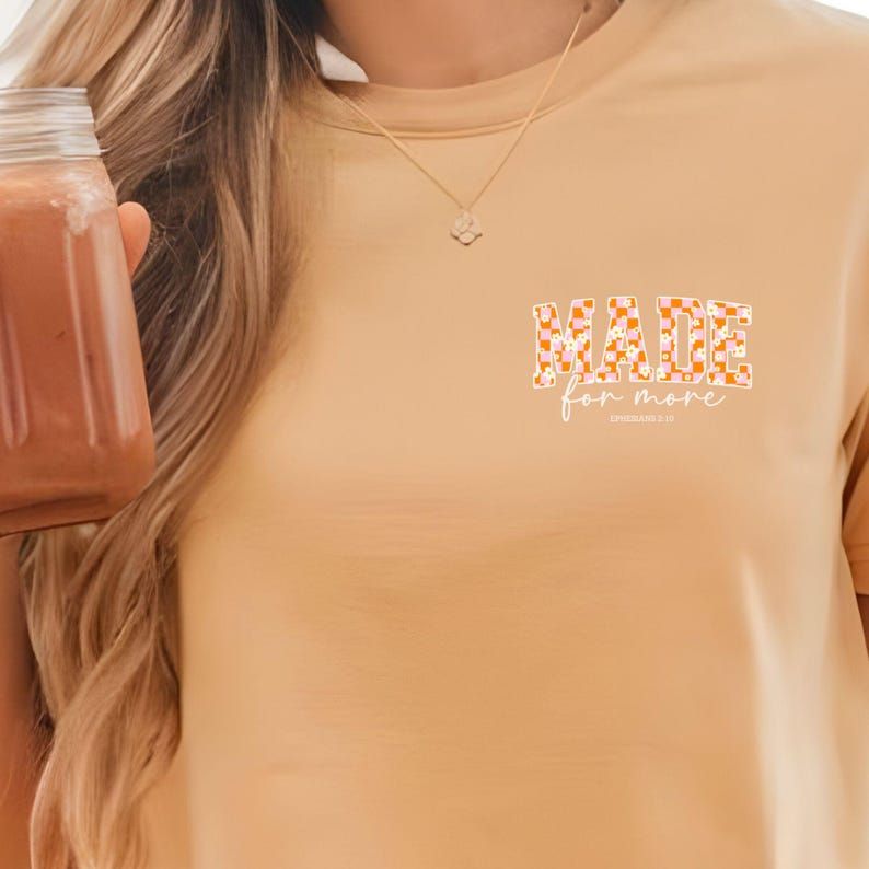 Puede incluir: Una camiseta de color beige con las palabras "MADE for more" en blanco. Las palabras est&aacute;n rodeadas de un patr&oacute;n a cuadros en naranja y rosa.