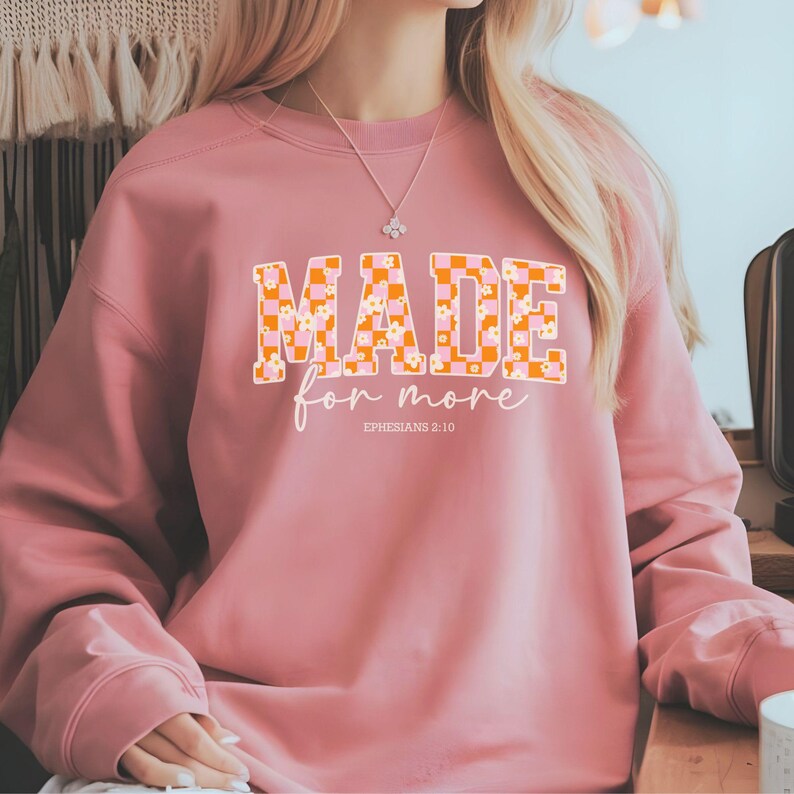 Puede incluir: Una sudadera rosa con un patr&oacute;n a cuadros y detalles florales. El texto "MADE for more" est&aacute; impreso en la parte delantera de la sudadera, con la referencia b&iacute;blica "EPHESIANS 2:10" debajo.