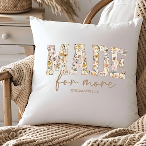 May include: White throw pillow with a floral design that spells out the word "MADE" with the text "for more" and "EPHESIANS 2:10" underneath.