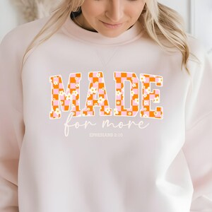 Puede incluir: Una sudadera blanca con el texto "MADE for more" en letras a cuadros naranja y rosa con flores blancas. El texto "EPHESIANS 2:10" est&aacute; impreso debajo del texto principal.