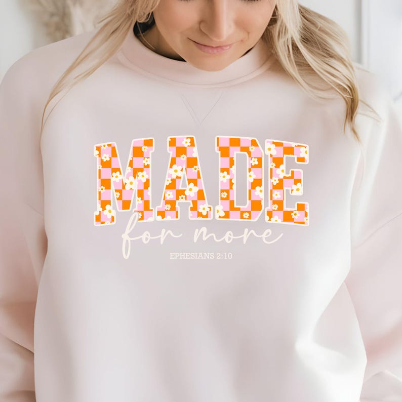 Puede incluir: Una sudadera blanca con el texto "MADE for more" en letras a cuadros naranja y rosa con flores blancas. El texto "EPHESIANS 2:10" est&aacute; impreso debajo del texto principal.