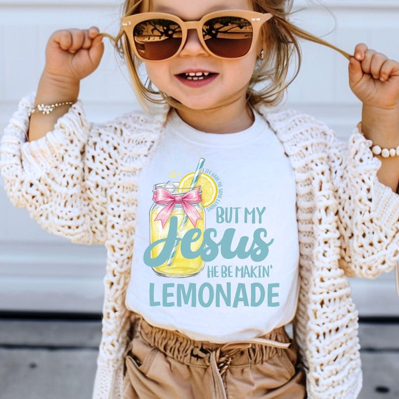 May include: White t-shirt with the text "BUT MY Jesus HE BE MAKIN' LEMONADE" in blue and yellow. The graphic includes a jar of lemonade with a pink bow and a lemon slice. The shirt is worn by a child wearing sunglasses.