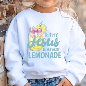 May include: White sweatshirt featuring a graphic of a lemonade jar with a pink bow and the text "BUT MY Jesus HE BE MAKIN' LEMONADE" in blue and yellow. The design includes a lemon slice and a straw.