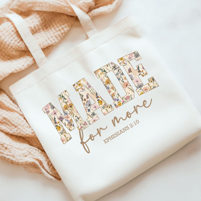 May include: White canvas tote bag with a floral design that spells out the word "MADE" and the text "for more" with the scripture reference "EPHESIANS 2:10" underneath.