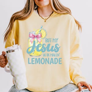 May include: Yellow sweatshirt with the text "BUT MY Jesus HE BE MAKIN' LEMONADE" and a graphic of a lemonade jar with a pink bow. The sweatshirt has a crew neck and long sleeves.