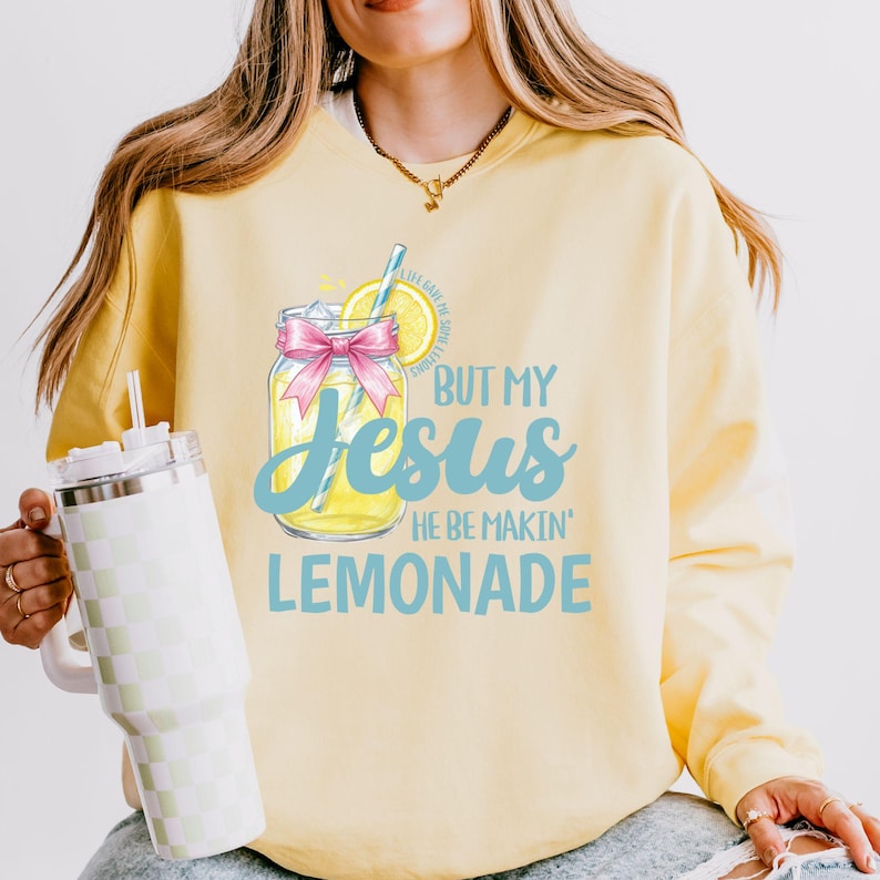 May include: Yellow sweatshirt with the text "BUT MY Jesus HE BE MAKIN' LEMONADE" and a graphic of a lemonade jar with a pink bow. The sweatshirt has a crew neck and long sleeves.