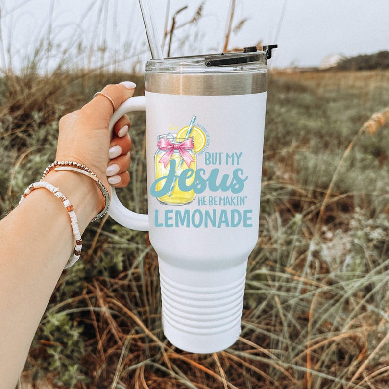 May include: White insulated tumbler with a handle and a silver lid. The tumbler features the text "BUT MY Jesus HE BE MAKIN' LEMONADE" with a graphic of a lemonade drink. A straw is visible.