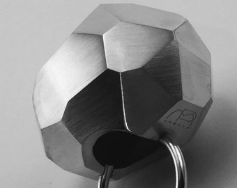 May include: A faceted, geometric silver-toned pendant with a circular ring. The pendant has a brushed metal finish and a dark interior. The letters "NB JEWELS" are engraved on the side. The design is modern and minimalist.