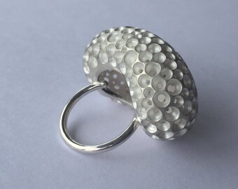 May include: A silver ring with a unique design. The ring features a large, textured, oval-shaped centerpiece covered in small, circular holes. The band is a simple, smooth silver loop. The ring is set against a plain, light background.